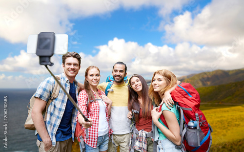 travel, tourism, hike and technology concept - group of smiling friends with backpacks taking picture by smartphone on selfie stick over big sur coast of california background