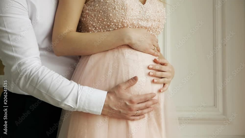 man husband embracing belly of his pregnant wife in dress standing next to window man husband embracing belly of his pregnant wife in dress standing next to window
