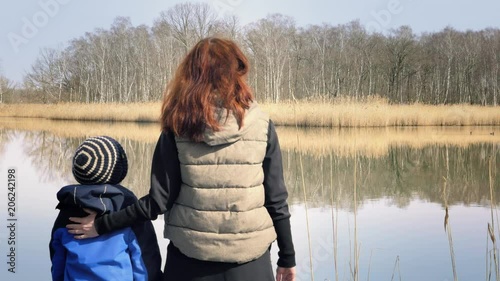 Woman and small boy are standing on small wooden pier at beautiful pond and enjoying countryside. Red-haired mother and son are enjoying nice lake. Sunny spring day, family values, happy atmosphere