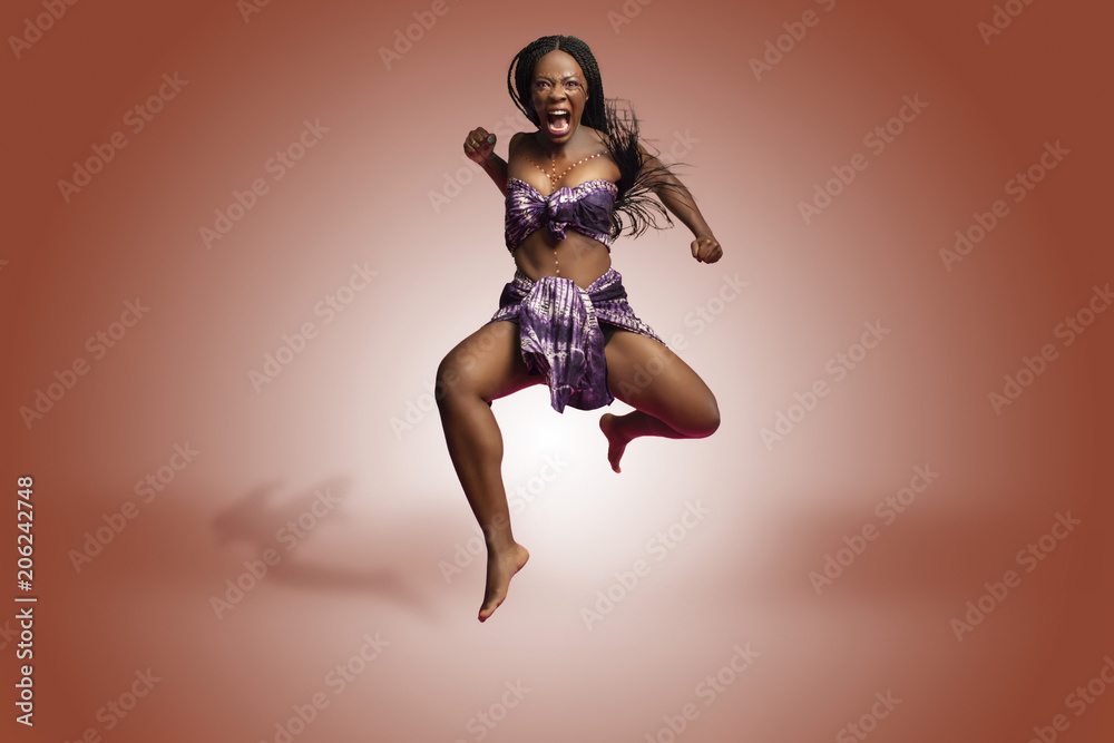 Beautiful African Black girl wearing traditional colorful African outfit does a dramatic aggressive leaping dance move against a colorful brown background