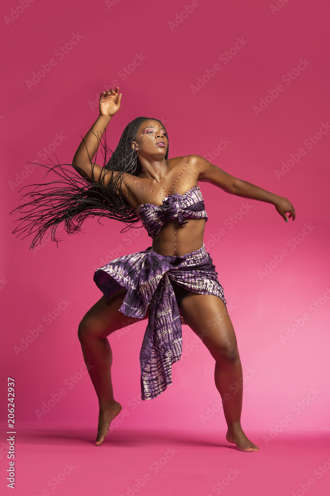 Beautiful African Black girl wearing traditional colorful African outfit does a dramatic dance move against a colorful pink background