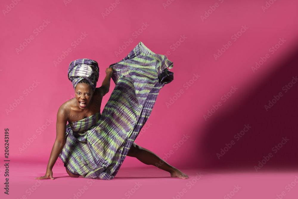 Beautiful African Black girl wearing traditional colorful African outfit does a dramatic dance move against a colorful pink background