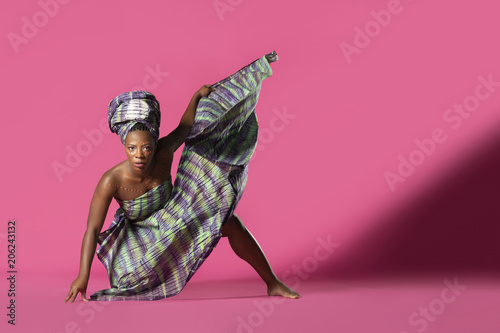 Beautiful African Black girl wearing traditional colorful African outfit does a dramatic dance move against a colorful pink background
