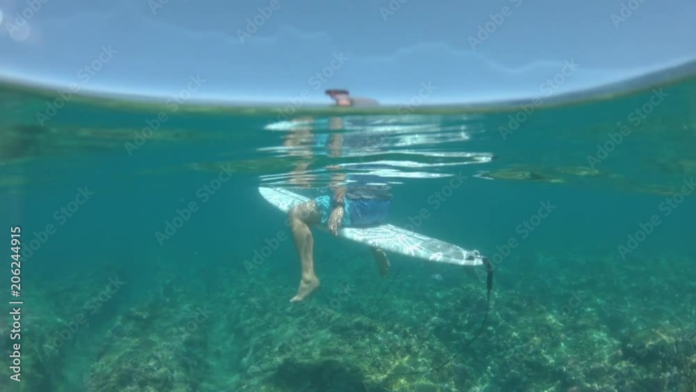 SLOW MOTION, HALF-UNDERWATER: Patient male surfer sits on his surfboard and waits for large waves to approach the shore. Surfboarder sits on his board in the turquoise ocean and looks around seascape. SLOW MOTION, HALF-UNDERWATER: Patient male surfer sits on his surfboard and waits for large waves to approach the shore. Surfboarder sits on his board in the turquoise ocean and looks around seascape.
