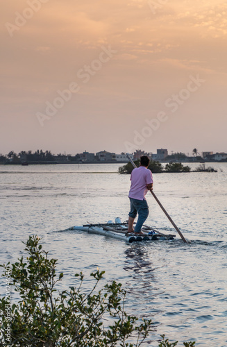 A man poles a raft out into the wetlands. It is late in the afternoon close to sunset. Small islands are in the wetlands. A distance shore can be seen. 