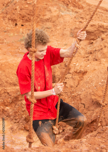 A young man splattered with mud climbing out of a pit using a rope during a mud run obstacle course