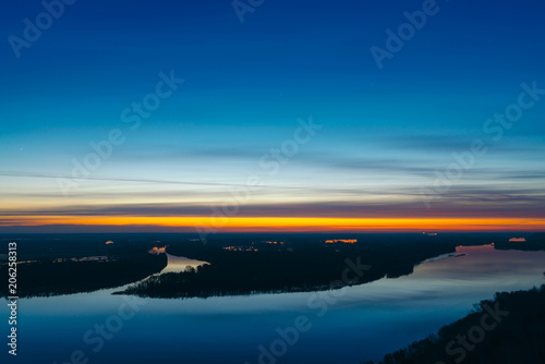 Beautiful river with big island with trees under predawn sky. Bright yellow stripe in picturesque cloudy sky. Early blue sky reflected in water. Colorful morning atmospheric image of majestic nature. © Daniil