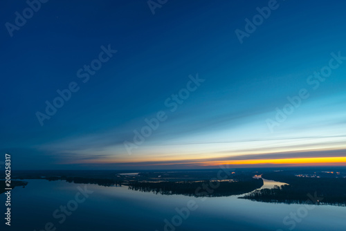 Beautiful river with big island with trees under predawn sky. Bright yellow stripe in picturesque cloudy sky. Early blue sky reflected in water. Colorful morning atmospheric image of majestic nature.