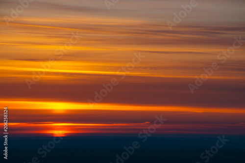 Big red sun circle rises out from behind dark horizon on background varicolored clouds of warmly shades. Beautiful background of dawn on picturesque cloud sky.
