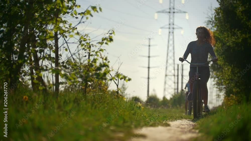 Two brunette cyclists smoothly pass along the path on the background of the blue sky and green trees Two brunette cyclists smoothly pass along the path on the background of the blue sky and green trees