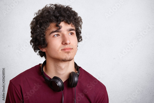Portrait of serious pensive curly male teenager with curly hair, uses modern headphones for listening favourite music, looks pensively aside, isolated on white background with blank copy space
