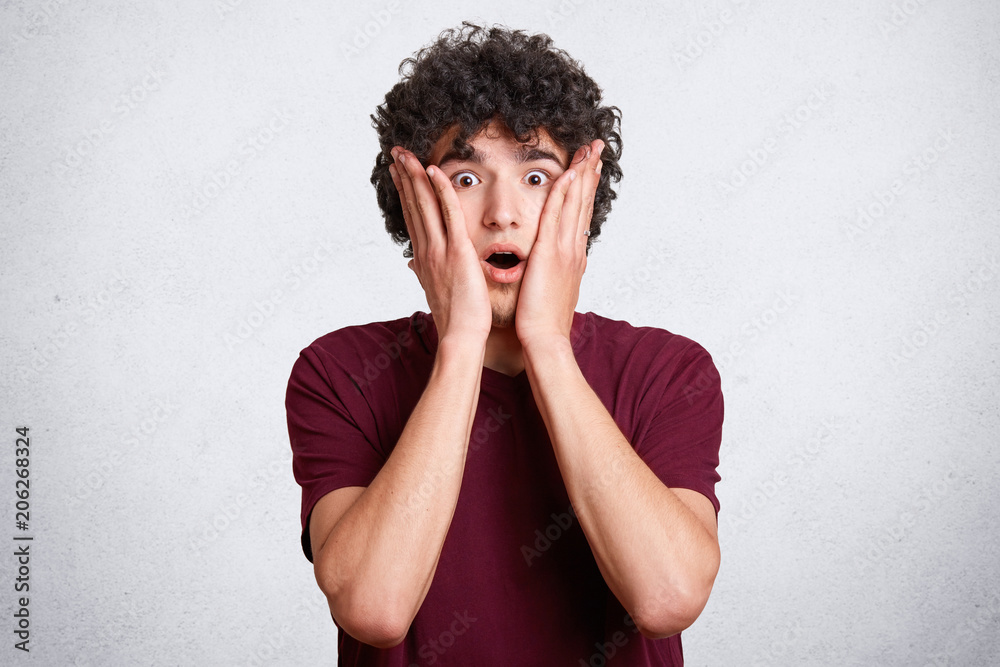 Portrait of handsome stunned male keeps hands on cheeks, jaw dropped, being speechless, surprised to hear shocking news, expresses surprisement, isolated on white concrete wall. Emotions concept Portrait of handsome stunned male keeps hands on cheeks, jaw dropped, being speechless, surprised to hear shocking news, expresses surprisement, isolated on white concrete wall. Emotions concept