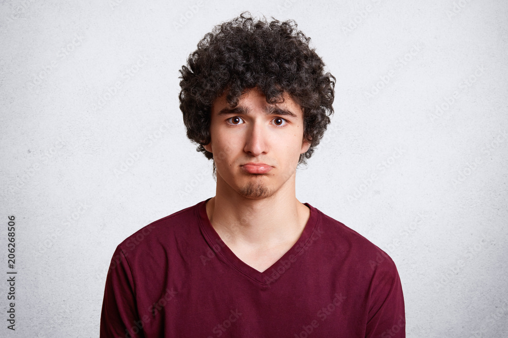 Headshot of upset attractive male curves lower lip, being dissatisfied with negative news, being sad and disappointed, poses against white concrete wall. Gloomy teenager discontent with something