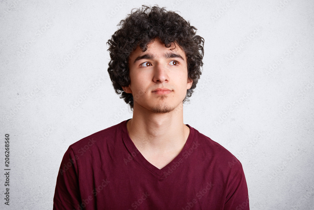 Obraz premium Close up shot of handsome curly teenager man looks pensively upwards, being deep in thoughts, ponders about future plans, isolated on white studio background. People, youth and lifestyle concept