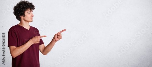 Horizontal portrait of handsome curly teenager has crisp dark hair, wears casual clothing, stands sideways and indicate with fore fingers at blank copy space for your promotional text or advertisement photo