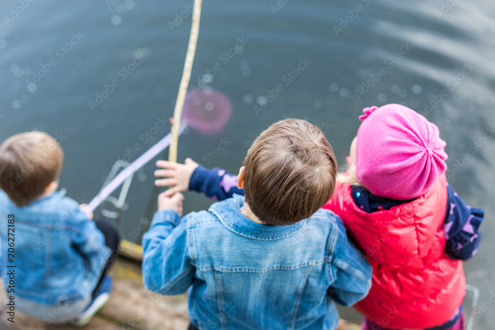 Three friends play fishing on wooden pier near pond. Two toddler