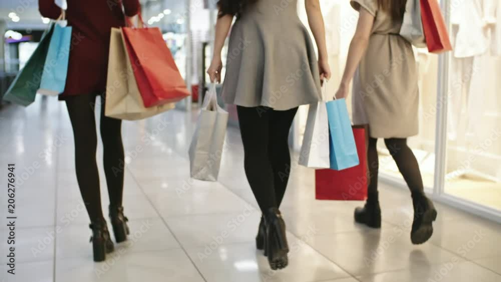 Tilt up shot of three young women carrying paper bags with purchases and looking into display windows when spending time together in shopping mall