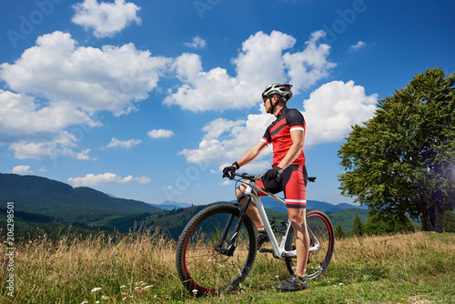 Young athletic tourist biker standing with bike on grassy valley enjoying beautiful view of distant Carpathian mountains on bright sunny day. Active lifestyle and extreme sport concept.
