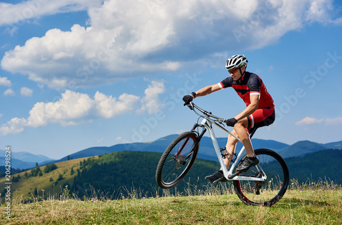 Young athletic sportsman tourist biker in professional sportswear turning on one wheel of bike on mountain hill on bright blue sky background. Active lifestyle and extreme sport concept. © anatoliy_gleb