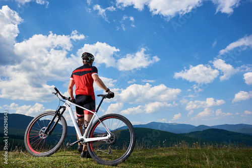 Back view of young athletic professional tourist biker standing at bike on top of hill, enjoying beautiful view of distant mountains on bright summer day. Active lifestyle and extreme sport concept.