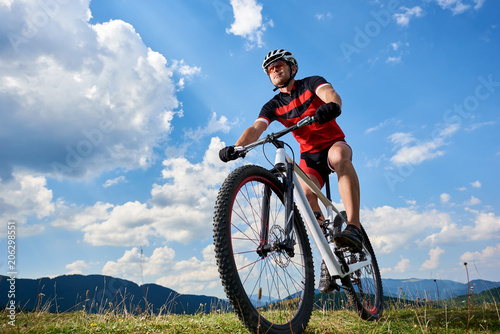 Close-up front view of young athletic sportsman cyclist in professional sportswear riding a bike on distant mountains and bright blue summer sky background. Active lifestyle and extreme sport concept.