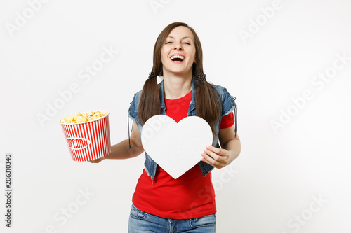 Portrait of young laughing beautiful woman in casual clothes watching movie film, holding bucket of popcorn and white heart with copy space isolated on white background. Emotions in cinema concept. photo