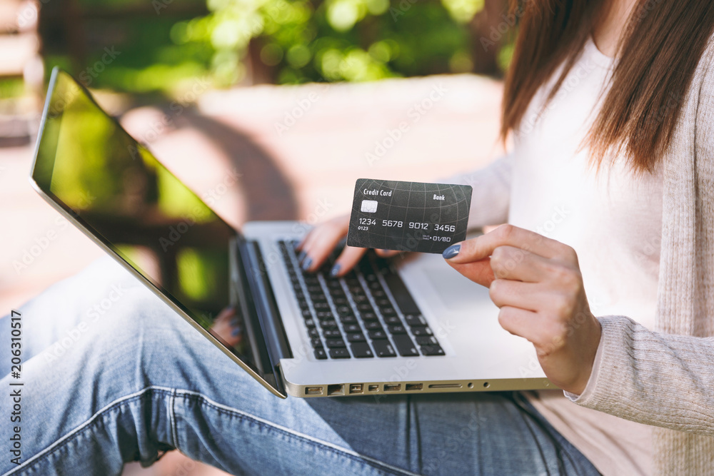 Naklejka premium Close up female hands on keyboard. Woman sitting on bench holding credit card, working on modern laptop pc computer in city park in street outdoors. Mobile Office. Online shopping, business concept.