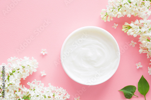 Opened white jar of natural herbal cream for women on pastel pink background. Beautiful branches of white lilac and petals. Care about clean and soft face, hands, legs and body skin. Fresh flowers.