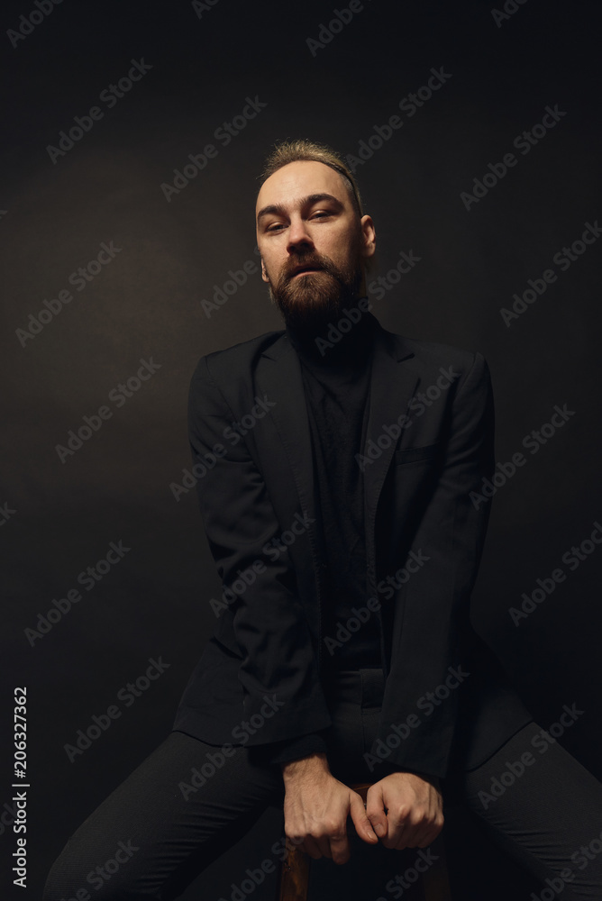 Feeling free and comfortable of his style. Handsome man in black jacket looking in camera while sitting on a chair. Theatre and emotions