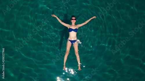 A beautiful girl in a blue bathing suit lies in the sea wearing sunglasses, top view, the copter flies up. The glare of the sun is reflected in the water. photo