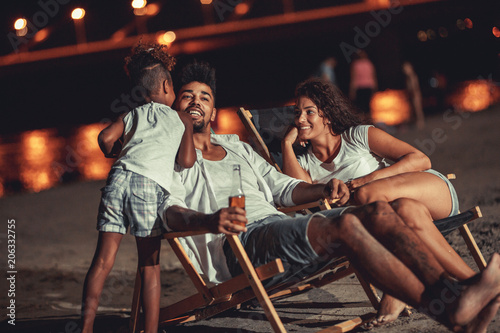 Young mixed race family sitting and relaxing at the beach on beautiful summer night.They sitting on sun beds and drink beer.