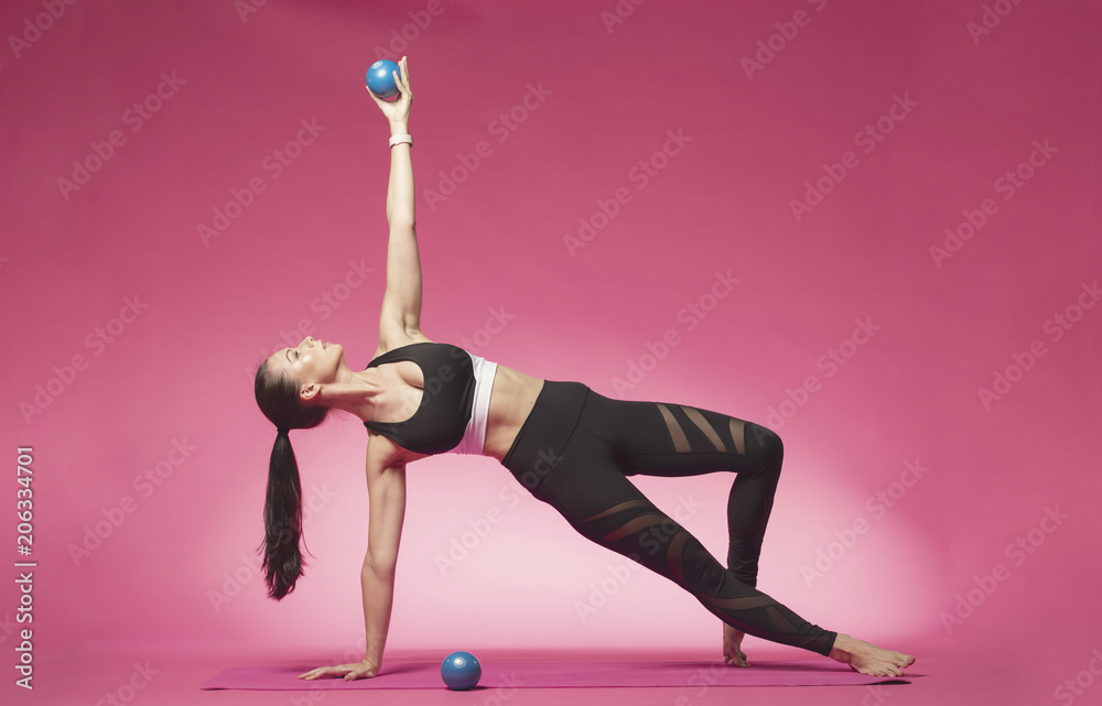 Long haired beautiful pilates or yoga athlete does a graceful pose with blue exercise balls while wearing a tight sports outfit against a pink background in a studio