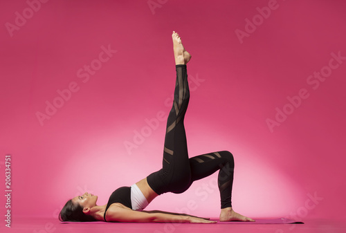 Long haired beautiful pilates or yoga athlete does a graceful pose while wearing a tight sports outfit against a pink background in a studio photo