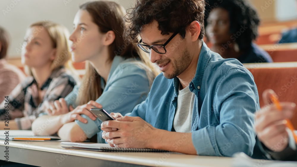Hispanic Man Uses Smartphone While Being on a Lecture in the Classroom. Lecture Hall Filled with Students Studying. Young People at University.