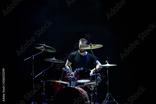 Drummer in a cap and headphones plays drums at a concert under white light in a smoke