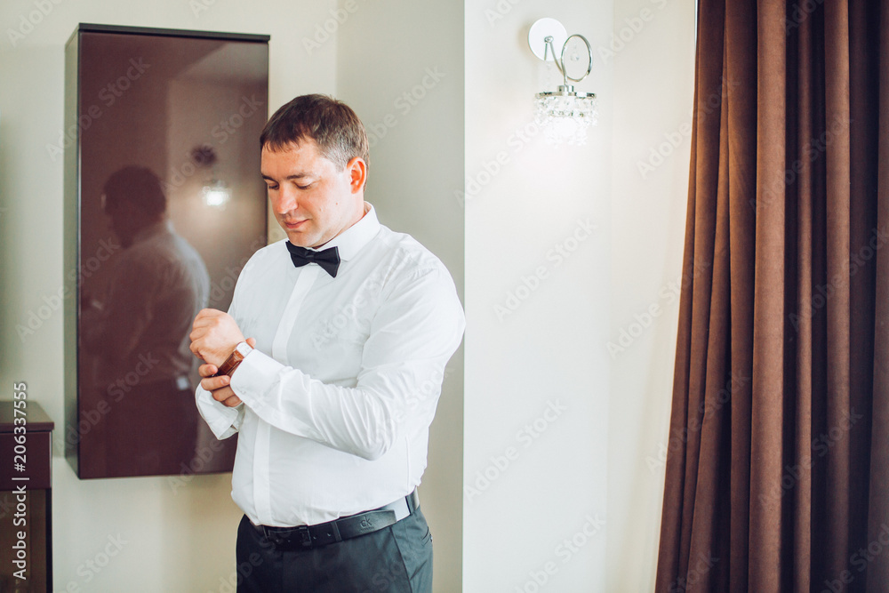 Fototapeta premium Stylish handsome groom posing while groomsman putting on shirt and tying bow, helping him to get ready in the morning. wedding preparation. 