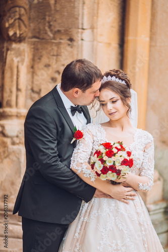 Beautiful fairytale newlywed couple hugging near old medieval castle. Wedding day for adorable newlyweds couple. Man and woomen hug and kiss aech other near old ancient door. photo