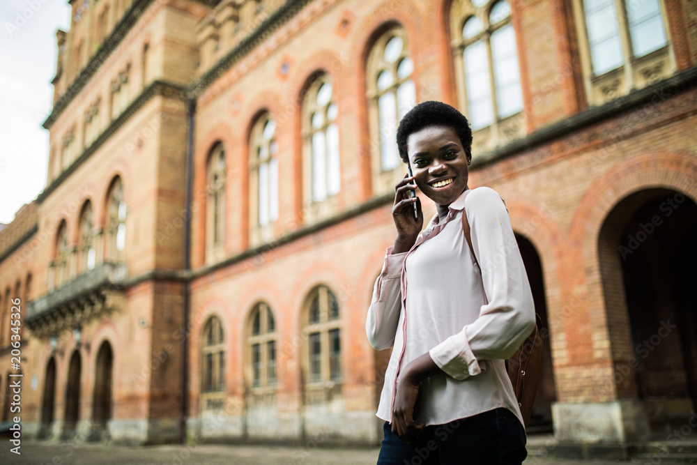 Fototapeta premium Portrait of pretty black woman in urban background talking on phone. Young student talking on phone at campus