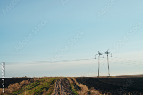 Atmospheric landscape with power lines in green field with dirt road under blue sky. Background image of electric pillars with copy space. Wires of high voltage above ground. Electricity industry.
