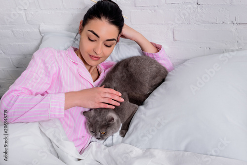 portrait of woman petting britain shorthair cat while resting in bed at home
