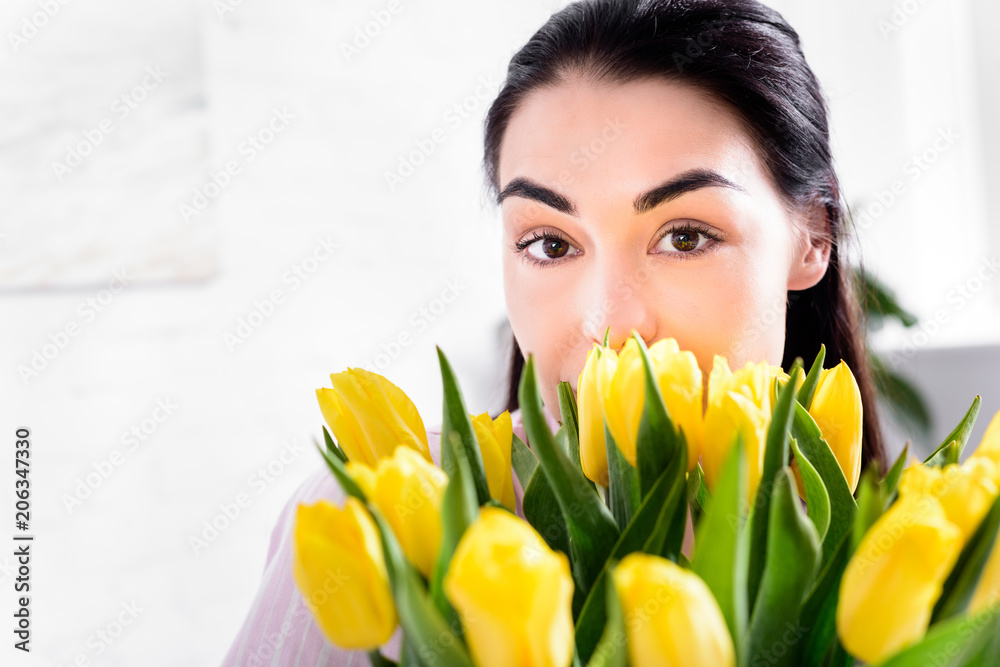 partial view of woman with bouquet of yellow tulips looking at camera at home partial view of woman with bouquet of yellow tulips looking at camera at home
