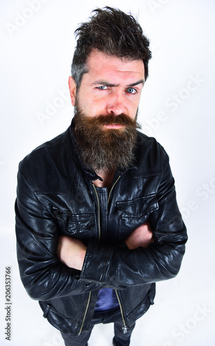 Man with beard and mustache on strict face looking at camera. Macho wears leather jacket, white background. Hipster looks serious while posing in stylish outfit. Menswear and fashion concept.