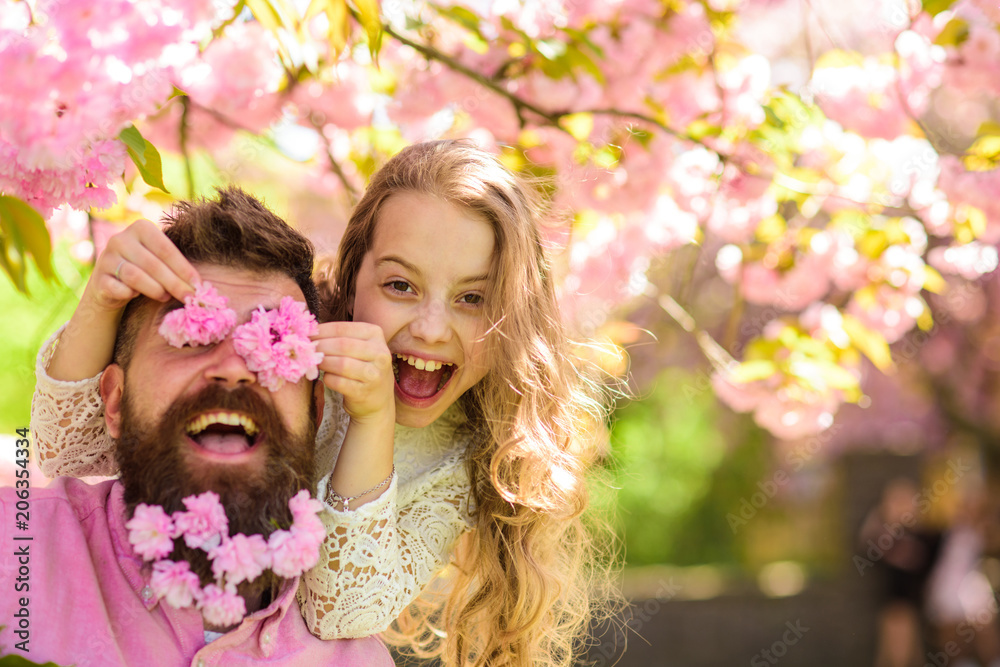 Fototapeta premium Dziecko i mężczyzna z delikatnymi różowymi kwiatami w brodzie. Dziewczyna z tatą w pobliżu kwiatów sakura na wiosenny dzień. Ojciec i córka na szczęśliwej twarzy bawią się kwiatami jako okulary, tło sakura. Pojęcie czasu dla rodziny
