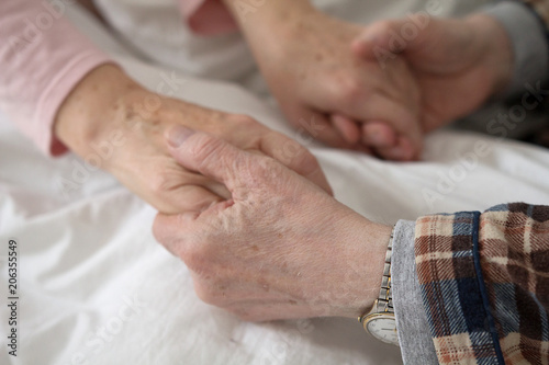 Aged man holding his wifes hands in bed on white bed sheet. Hands of old man with wrist watch holding old womans hands.