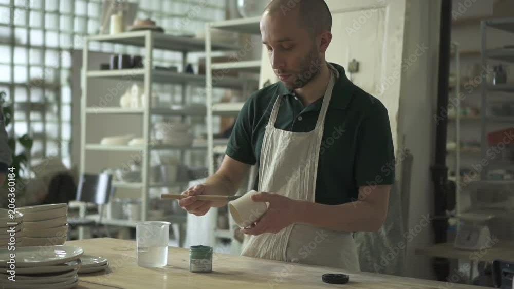 Young artist is painting on earthenware in pottery studio. Talented man draws on mug with brush while standing at table indoors. Professional covers white product with color using fluffy tassel Young artist is painting on earthenware in pottery studio. Talented man draws on mug with brush while standing at table indoors. Professional covers white product with color using fluffy tassel