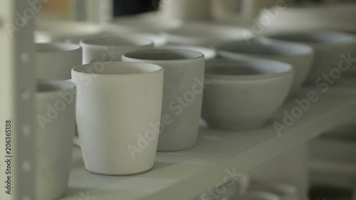 Close up of bowls and mugs on the shelf in ceramic studio. White dishes are stanging on stack after applying of enamel, drying and waiting for baking in kiln. This makes creatures prepared to useage. photo