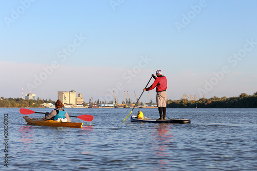 Water tourists is paddling on SUP (Stand up paddle board) near woman in kayak at Danube river on the background of port cranes