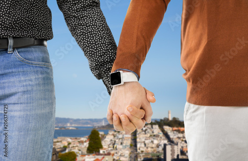 same-sex relationships, lgbt and technology concept - close up of male gay couple holding hands with smartwatch over san francisco city background