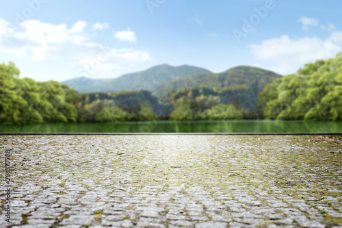 Empty square made of stone cubes against the blurred background of a green park with a lake and mountains and sky in the background.