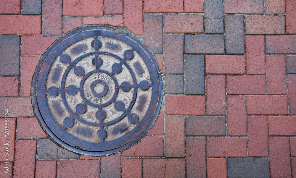 Close up photo of a storm sewer manhole cover on a brick sidewalk.